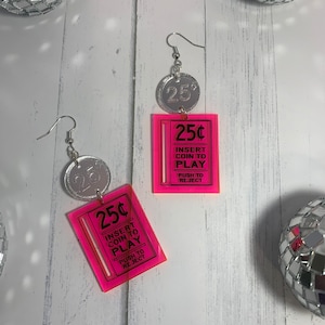 May include: A pair of pink acrylic earrings with silver accents. Each earring features a pink rectangle with the text "25¢ INSERT COIN TO PLAY PUSH TO REJECT" and a silver coin with the number "25" on it.