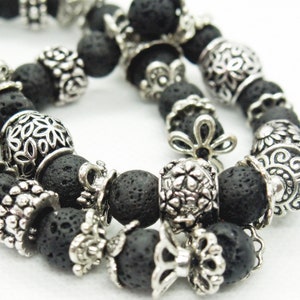 Könnte beinhalten: Zwei schwarze Lavastein-Armbänder mit silberfarbenen Zierperlen. Die Perlen weisen florale, filigrane und strukturierte Designs auf. Die Armbänder sind vor einem weißen Hintergrund platziert, was den Kontrast hervorhebt.