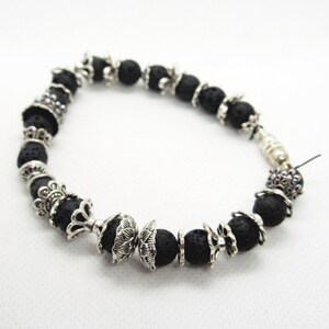 Könnte beinhalten: Ein Armband aus schwarzem Lavastein mit silberfarbenen Zierperlen. Das Armband besteht aus runden, porösen schwarzen Perlen, die mit verzierten silberfarbenen Metallakzenten durchsetzt sind. Der Verschluss ist ebenfalls silberfarben.