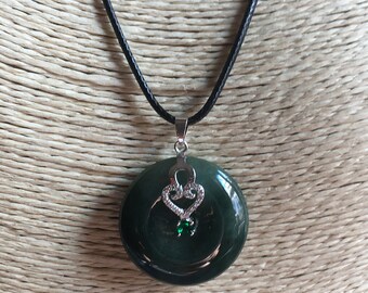 May include: A dark green jade pendant necklace with a silver heart charm. The pendant is round and has a smooth, polished surface. The necklace is made of black cord.