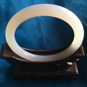 Puede incluir: Un brazalete de jade blanco roto y liso. El brazalete circular se exhibe sobre un soporte de madera marrón oscuro. El jade tiene un aspecto pulido y translúcido.