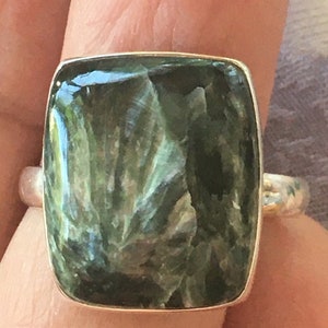 Puede incluir: Un anillo de plata con una gran piedra preciosa verde de corte cuadrado. La piedra tiene un patrón arremolinado distintivo y está engastada en una banda de plata pulida. El diseño del anillo es simple y elegante, adecuado para el uso diario o ocasiones especiales.