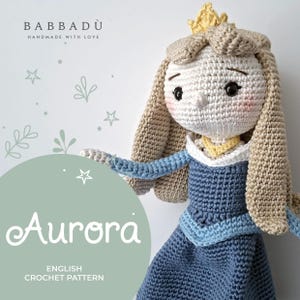 Può includere: Una bambola realizzata all'uncinetto a mano che raffigura un personaggio con lunghi capelli biondi, una piccola corona gialla e un vestito blu con colletto bianco. L'immagine include il testo BABBADU HANDMADE WITH LOVE e Aurora ENGLISH CROCHET PATTERN.