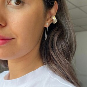 May include: A woman wearing a white t-shirt with a silver chain earring with a star charm and a silver hoop earring with a textured design.