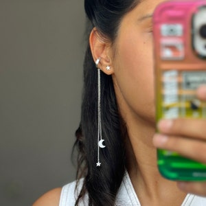 May include: A silver ear cuff with a dangling chain featuring a crescent moon and two stars. The chain is attached to the ear cuff with a small silver ring.