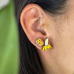 Peut inclure: Une paire de boucles d'oreilles en forme de banane jaune et marron avec un visage souriant sur une banane. Les boucles d'oreilles sont portées par une personne aux cheveux foncés.