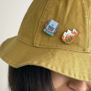 Può includere: Una persona indossa un cappello a secchiello giallo senape con due spille smaltate. Una spilla è un'illustrazione blu e bianca di uno zaino, l'altra spilla è un'illustrazione marrone di montagne con alberi verdi.