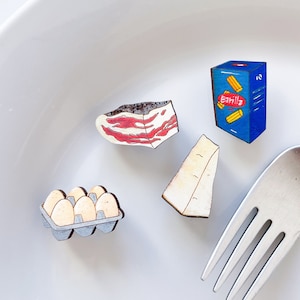 May include: A white plate with a fork, a carton of pasta, a carton of eggs, a piece of cheese, and a piece of bacon. The pasta carton is blue with the word "Barilla" on it.
