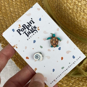 Può includere: Un biglietto bianco con il testo "Pop'n Tags Design" e un indirizzo di sito web. Il biglietto ha due piccole spille di legno, una di una tartaruga marina e l'altra di una conchiglia.