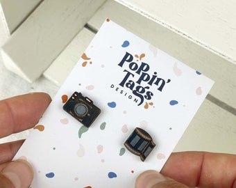 Op de afbeelding: Twee houten pinnen op een witte kaart met gekleurde stippen. De pinnen zijn gevormd als een vintage camera en een filmrol. De kaart heeft de tekst "Pop'n' Tags Design" en "poppintagsdesign.com".
