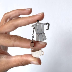 Puede incluir: Una cafetera y una taza de madera cuelgan de una cadena. La cafetera está pintada de gris y tiene un asa negra. La taza está pintada de blanco con una mancha de café marrón en el interior.