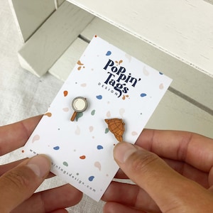 Könnte beinhalten: Zwei Holz-Pins auf einer weißen Karte mit einem bunten Konfetti-Muster. Die Pins sind in Form einer Lupe und einer Muschel geformt. Die Karte trägt den Text "Popin' Tags Design".