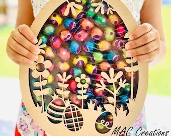 May include: A brown wooden Easter egg-shaped craft with a cut-out design of flowers and grass. The craft is filled with colorful foil-wrapped chocolate eggs. The text "MAC Creations" is visible at the bottom of the image.