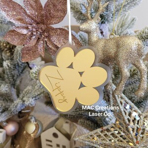 May include: Gold glitter paw print bauble with the name "Zippy" engraved on it. The bauble is hanging on a Christmas tree with other gold glitter decorations.