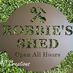 May include: A brown metal sign with the text "Robbie's Shed Open All Hours" and a crossed wrench and hammer design. The sign is surrounded by green foliage.