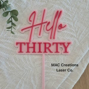 May include: A pink acrylic cake topper with the words "Hello THIRTY" in a script and block font. The topper has a long, thin stem for insertion into a cake. The text is in a darker pink colour.