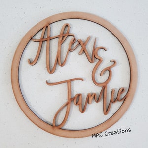 May include: A wooden circle with the names "Alex & Jamie" written in cursive script. The circle is brown and has a natural wood finish. The text "MAC Creations" is printed in the bottom right corner.