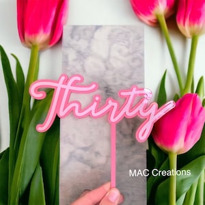 May include: A pink and light pink layered "Thirty" cake topper with a stem, held in a hand. The background includes pink tulips and a marble-patterned surface. The text "MAC Creations" is visible.