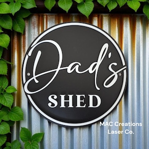 May include: A circular black sign with the words "Dad's SHED" in white script. The sign has a white border and is set against a corrugated metal backdrop. Green leaves surround the sign.
