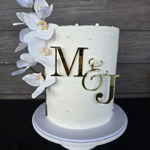 May include: A white cake decorated with white orchids and gold-coloured initials "M & J". The cake is on a white cake stand and has small white pearl-like decorations. The background is a wooden wall.