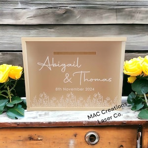 May include: A light brown wooden box with a slot on the top. The box is decorated with white lettering that says "Abigail & Thomas" and "8th November 2024". There are also white floral designs on the box. The box is sitting on a wooden surface. The text "MAC Creations Laser Co." is visible in the bottom right corner.