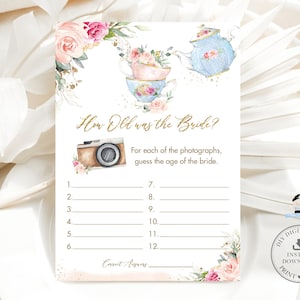 以下が含まれることがあります： 「How Old was the Bride?」の文字が金色の印刷可能なブライダルシャワーゲームカード。カードには、ティーカップ、ティーポット、カメラの水彩イラストが描かれており、ピンクとピーチの花柄のアクセントが付いています。