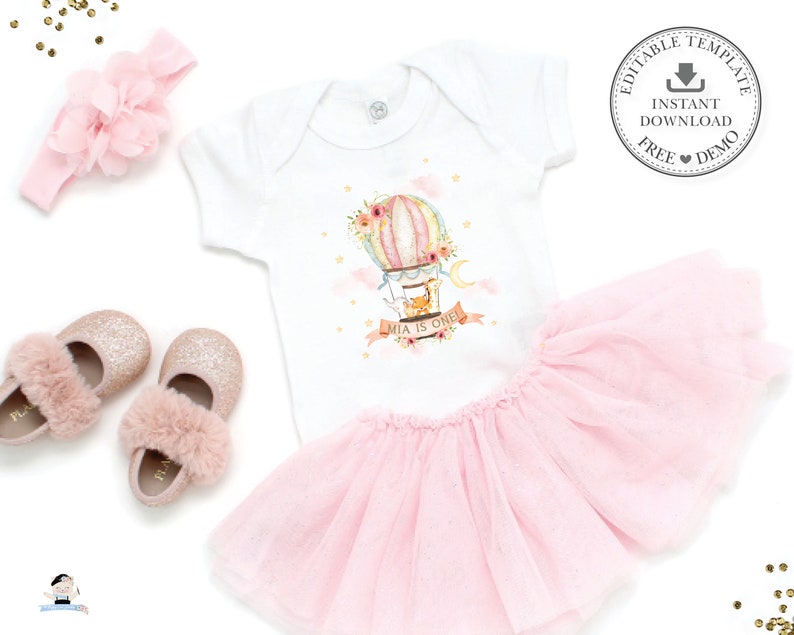 May include: A pink tutu skirt, a white onesie with a hot air balloon graphic and the text "Mia is one!" and a pink headband with a flower.