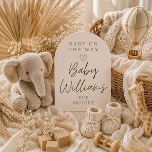 May include: An arched wooden sign with the text BABY ON THE WAY, a heart icon, Baby Williams, and DUE 08/20XX. The sign is surrounded by a plush gray elephant, a pair of cream knit baby booties, a wooden giraffe, a wooden rattle, and blocks.