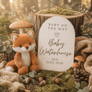May include: A plush orange and white fox toy sits next to an arched wooden sign that reads BABY ON THE WAY, a heart icon, Baby Waterhouse, and DUE JUNE 2026. The scene includes small mushrooms, acorns, and a pinecone on a mossy forest floor.