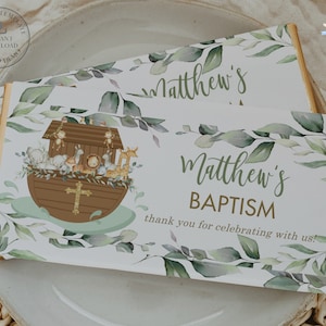 Puede incluir: Una barra de chocolate blanco de favor con un diseño de hojas verdes y el texto "Bautismo de Matthew" y "¡Gracias por celebrar con nosotros!" La barra presenta una ilustración de dibujos animados del Arca de Noé con animales.