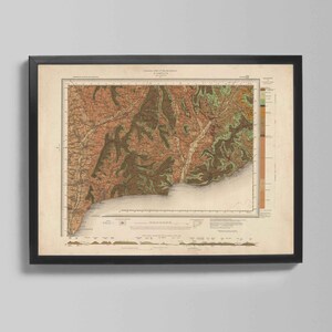 May include: Framed vintage map of a coastal region, featuring detailed topographical information. The map uses a palette of earthy tones, including browns, greens, and tans, with a black frame. The map is mounted on a neutral background.