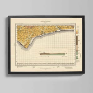 May include: Framed vintage map print featuring a detailed geological survey. The map displays a coastal region with yellow, brown, and green hues, set against a grid background. The print is encased in a black frame, suitable for home decor.