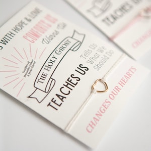 May include: White string bracelet with a gold heart charm. The bracelet has a card attached with the text "Teaches Us" and "Changes Our Hearts". The card also includes the text "The Holy Ghost" with a banner design.
