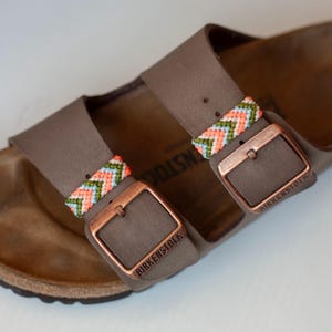 May include: Close-up of a pair of brown leather sandals with copper-colored buckles. The straps are adorned with colorful woven patterns in shades of orange, green, and blue. The brand name "BIRKENSTOCK" is visible on the buckles.