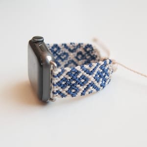 May include: A silver smartwatch with a woven band. The band features a geometric pattern in navy blue and cream colors. The watch is displayed on a white surface.