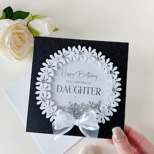 Peut inclure: Une carte noire avec une couronne de marguerites blanches avec des centres roses et des paillettes argent&eacute;es. La carte dit "Joyeux anniversaire &agrave; une fille tr&egrave;s sp&eacute;ciale".