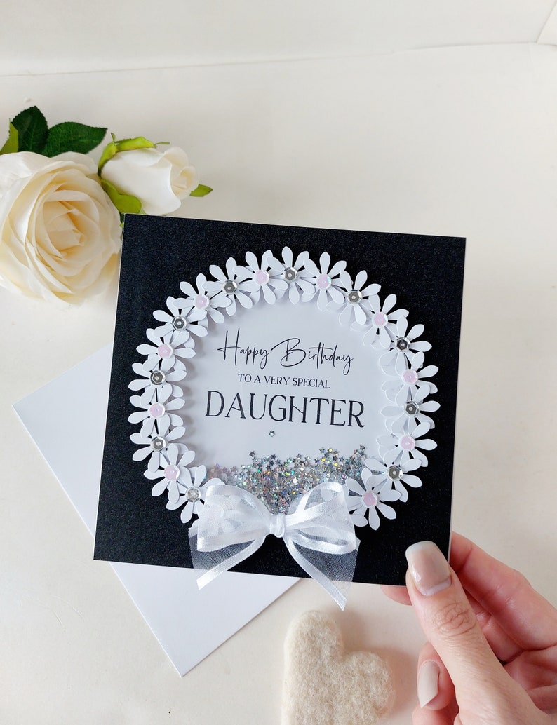 Peut inclure: Une carte noire avec une couronne de marguerites blanches avec des centres roses et des paillettes argent&eacute;es. La carte dit "Joyeux anniversaire &agrave; une fille tr&egrave;s sp&eacute;ciale".