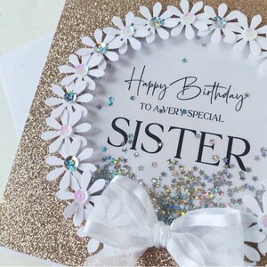 May include: A white card with a gold glitter background and a white daisy wreath with rhinestones. The card says "Happy Birthday to a very special Sister" with silver glitter stars scattered around the text.