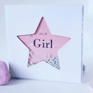 K&ouml;nnte beinhalten: Eine wei&szlig;e Karte mit einem rosafarbenen sternf&ouml;rmigen Fenster. Der Stern ist mit silbernem Glitzer gef&uuml;llt und der Text "It's a Girl" ist darin gedruckt.