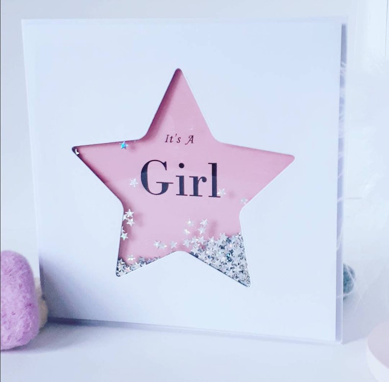 K&ouml;nnte beinhalten: Eine wei&szlig;e Karte mit einem rosafarbenen sternf&ouml;rmigen Fenster. Der Stern ist mit silbernem Glitzer gef&uuml;llt und der Text "It's a Girl" ist darin gedruckt.