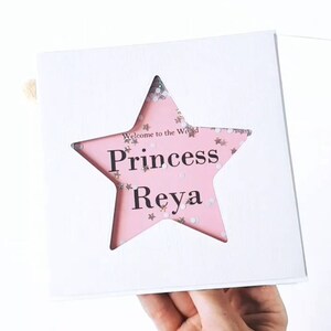 K&ouml;nnte beinhalten: Wei&szlig;e Karte mit einem rosafarbenen sternf&ouml;rmigen Fenster. Der Stern ist mit Glitzer gef&uuml;llt und der Text "Welcome to the World Princess  Reya" ist durch das Fenster sichtbar.