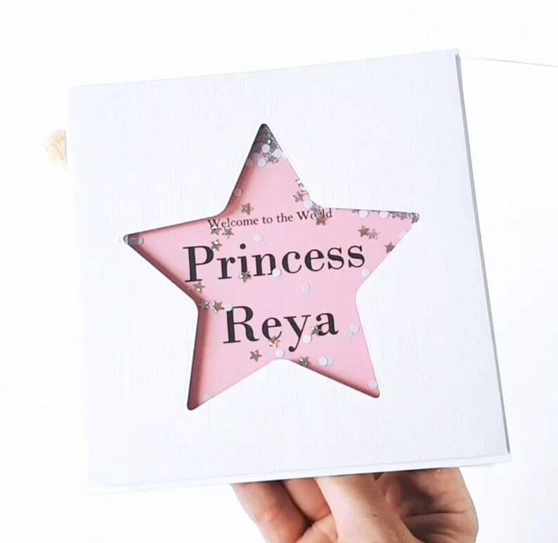 K&ouml;nnte beinhalten: Wei&szlig;e Karte mit einem rosafarbenen sternf&ouml;rmigen Fenster. Der Stern ist mit Glitzer gef&uuml;llt und der Text "Welcome to the World Princess  Reya" ist durch das Fenster sichtbar.
