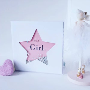 K&ouml;nnte beinhalten: Eine wei&szlig;e Karte mit einem ausgeschnittenen rosa Stern. Der Stern hat Glitzer im Inneren und die Worte "It's a Girl" sind darauf geschrieben.