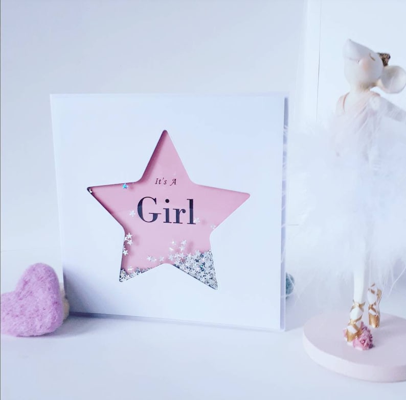 K&ouml;nnte beinhalten: Eine wei&szlig;e Karte mit einem ausgeschnittenen rosa Stern. Der Stern hat Glitzer im Inneren und die Worte "It's a Girl" sind darauf geschrieben.