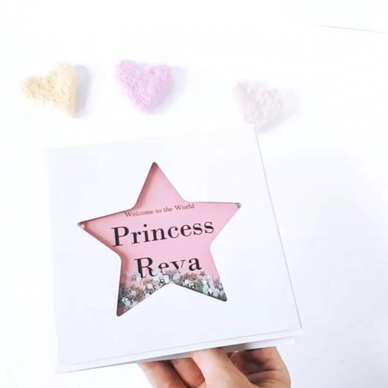 K&ouml;nnte beinhalten: Eine wei&szlig;e Karte mit einem ausgeschnittenen rosa Stern. Der Stern enth&auml;lt Glitzer und die Worte "Willkommen auf der Welt Prinzessin Reva" sind darauf gedruckt.