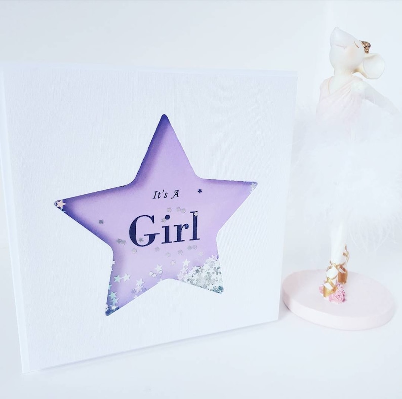 K&ouml;nnte beinhalten: Eine wei&szlig;e Karte mit einem ausgeschnittenen lila Stern. Der Stern hat Glitzer im Inneren und die Worte "It's A Girl" in schwarzem Text.
