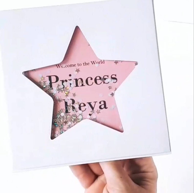 K&ouml;nnte beinhalten: Ein rosa sternf&ouml;rmiger Rahmen mit Glitzer im Inneren. Der Rahmen enth&auml;lt den Text "Welcome to the World Princess Reya" im Inneren.