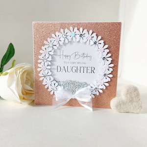 Peut inclure: Une carte blanche avec un fond de paillettes or rose. La carte a un cercle de papier blanc avec une couronne de marguerites blanches autour. La couronne est d&eacute;cor&eacute;e de strass roses et bleus. Le texte &agrave; l'int&eacute;rieur du cercle dit "Joyeux anniversaire &agrave; une fille tr&egrave;s sp&eacute;ciale".