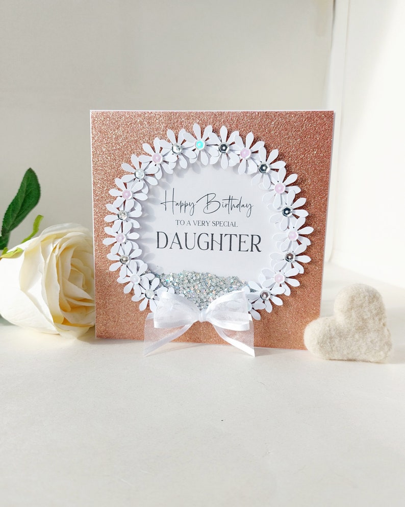 Peut inclure: Une carte blanche avec un fond de paillettes or rose. La carte a un cercle de papier blanc avec une couronne de marguerites blanches autour. La couronne est d&eacute;cor&eacute;e de strass roses et bleus. Le texte &agrave; l'int&eacute;rieur du cercle dit "Joyeux anniversaire &agrave; une fille tr&egrave;s sp&eacute;ciale".