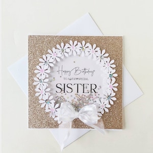 May include: A gold glitter card with a white daisy wreath and a white bow. The card says "Happy Birthday to a very special Sister".
